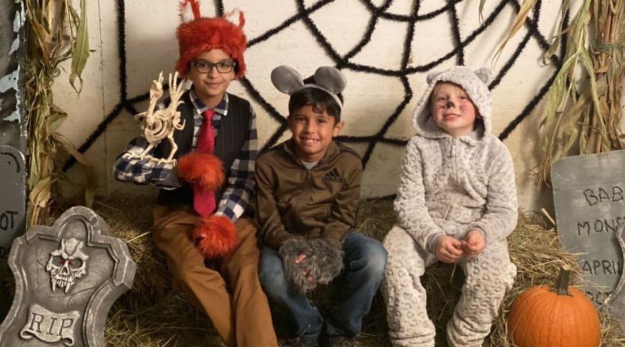 Three young kids in cute Halloween costumes sit on hay bales decorated with pumpkins and tombstones with dried corn stalks and a big black spider web hanging on the wall behind them at the Not So Haunted Stony Kill Spooktacular in Wappingers Falls